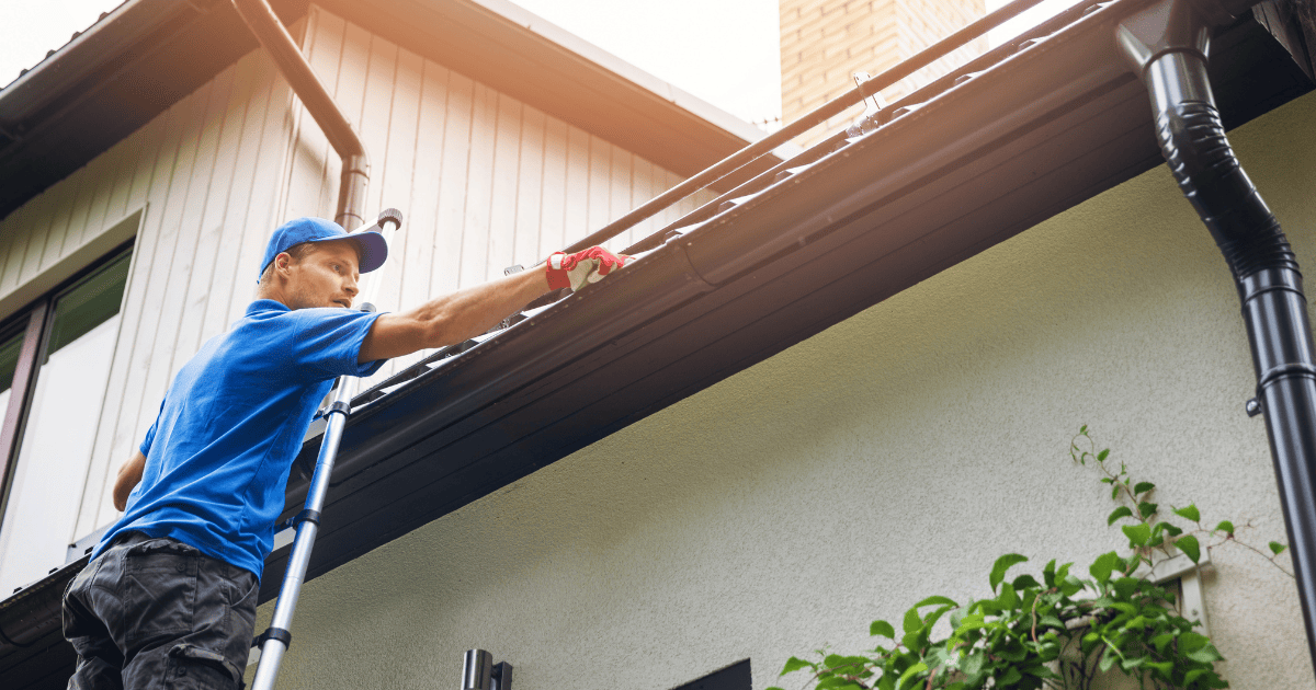 a handyman on a ladder reaching into a home's gutters to clean it