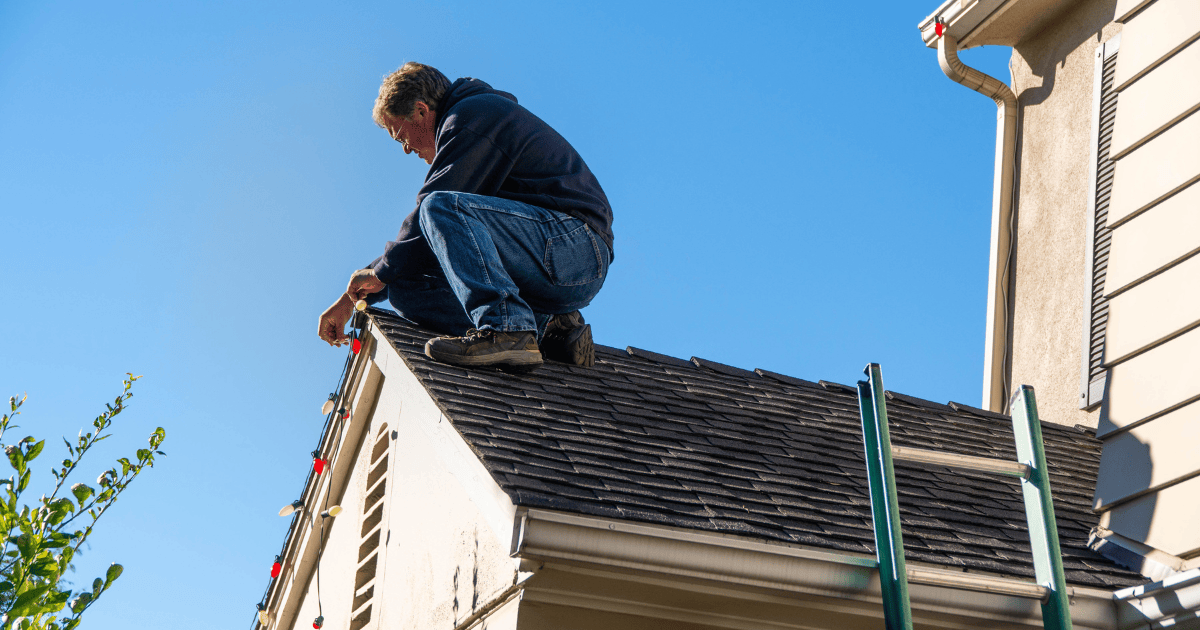 a handyman sitting on the peak of a roof hanging Christmas lights