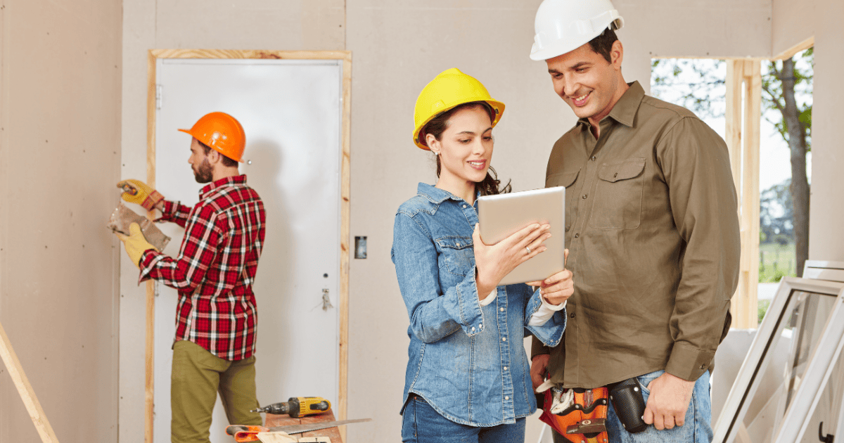 a contractor installing drywall while a man and woman wearing hard hats look at room remodel plans on a tablet
